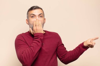 Yetişkin, yakışıklı bir adam kendini mutlu, şaşkın ve şaşırmış hissediyor. Ağzını eliyle kapatıyor ve yanal kopya alanını işaret ediyor.