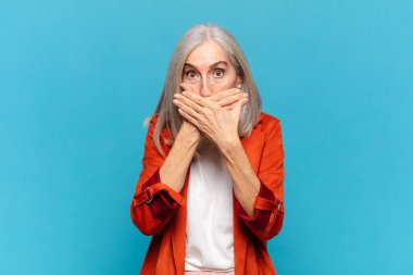 Orta yaşlı bir kadın şok olmuş, şaşırmış bir ifadeyle ağzını kapatıyor, sır saklıyor ya da 