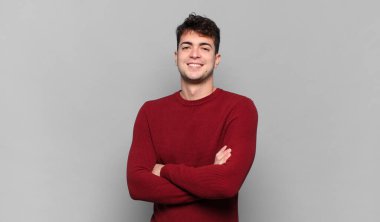 Genç adam kameraya çapraz kollarla gülümsüyor ve mutlu, kendinden emin, tatmin olmuş bir ifade, yan bakış.