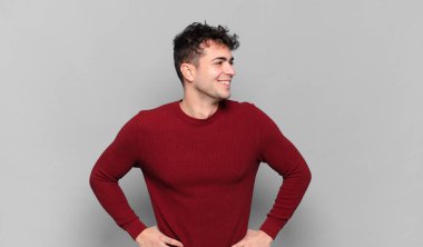 Genç adam mutlu, neşeli ve kendinden emin görünüyor, gururla gülümsüyor ve iki elini de kalçasında istiyor.