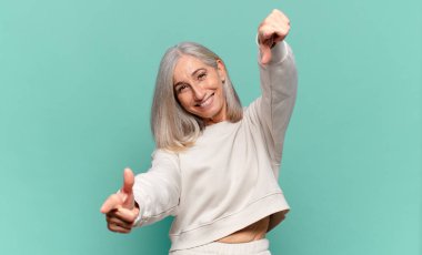 Orta yaşlı bir kadın mutlu ve kendinden emin, kamerayı iki eliyle gösteriyor ve gülüyor, seni seçiyor.