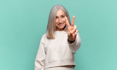 Orta yaşlı kadın gülümsüyor ve mutlu görünüyor, kaygısız ve pozitif, zafer ya da barış işareti yapıyor.