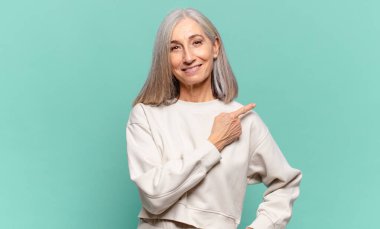 Orta yaşlı kadın neşeli bir şekilde gülümsüyor, mutlu hissediyor ve yan ve yukarı doğru işaret ediyor, fotokopi uzayındaki nesneleri gösteriyor.