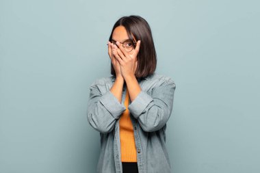 Latin kökenli genç bir kadın korkmuş ya da utanmış hissediyor. Gözleri yarı kapalı ya da elleri kapalı gözetliyor.