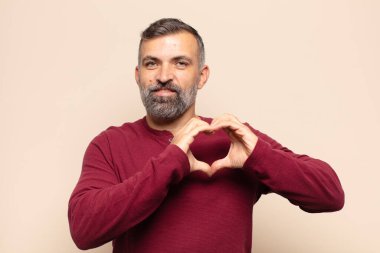 Yetişkin, yakışıklı bir adam gülümsüyor ve mutlu, sevimli, romantik ve aşık, iki eliyle kalbi şekillendiriyor.