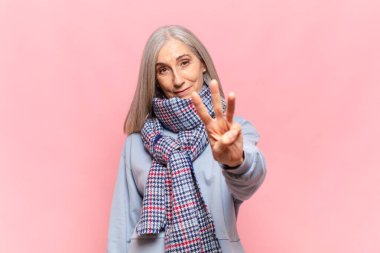 Orta yaşlı bir kadın gülümsüyor ve arkadaş canlısı görünüyor. Üç ya da üç numarayı gösteriyor. Elleri önde, geri sayıyor.