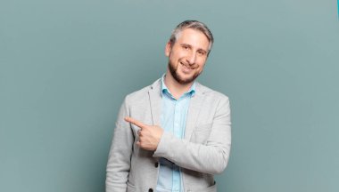 Yetişkin iş adamı neşeli bir şekilde gülümsüyor, mutlu hissediyor ve kenarı ve yukarıyı işaret ediyor, fotokopi alanında nesneler gösteriyor.