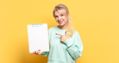 Sarışın kadın neşeyle gülümsüyor, mutlu hissediyor ve yan ve yukarı doğru işaret ediyor, fotokopi uzayındaki nesneleri gösteriyor.