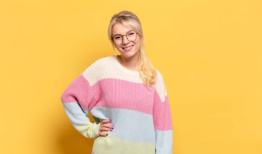 Sarışın kadın mutlu bir şekilde gülümsüyor, eli kalçasında ve kendinden emin, olumlu, gururlu ve arkadaş canlısı.