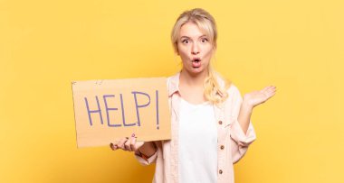 Sarışın kadın şaşırmış ve şoka uğramış görünüyor. Çenesi düşmüş ve eli açık bir objeyi tutuyor.