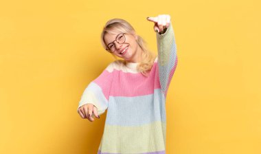 Sarışın kadın mutlu ve kendinden emin, kamerayı iki eliyle gösteriyor ve gülüyor, seni seçiyor.