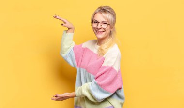 Sarışın kadın gülümsüyor, mutlu, pozitif ve tatmin olmuş hissediyor, fotokopi alanında nesne veya konsept tutuyor veya gösteriyor