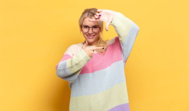 Sarışın kadın mutlu, arkadaş canlısı ve pozitif hissediyor, gülümsüyor ve elleriyle bir portre veya fotoğraf çerçevesi yapıyor.