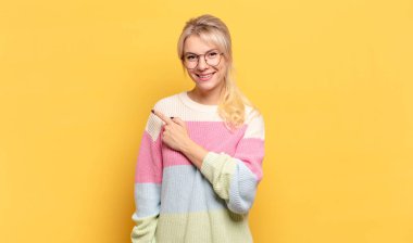 Sarışın kadın neşeyle gülümsüyor, mutlu hissediyor ve yan ve yukarı doğru işaret ediyor, fotokopi uzayındaki nesneleri gösteriyor.