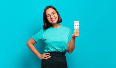 Genç İspanyol kadın mutlu bir şekilde gülümsüyor, eli kalçasında ve kendinden emin, olumlu, gururlu ve arkadaş canlısı bir tavırla.