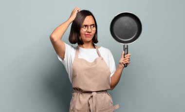 Genç İspanyol kadın şaşkın ve şaşkın hissediyor, kafasını kaşıyor ve yan tarafa bakıyor.