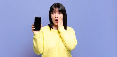 Artı boyunda güzel bir kadın şok olmuş ve korkmuş hissediyor, ağzı açık ve elleri yanaklarında korkmuş görünüyor.