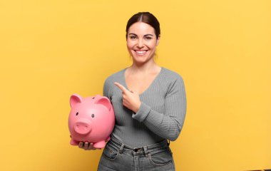 Genç, güzel bir kadın neşeli bir şekilde gülümsüyor, mutlu hissediyor ve kenarı ve yukarıyı işaret ediyor, kopyalama alanında nesneler gösteriyor.