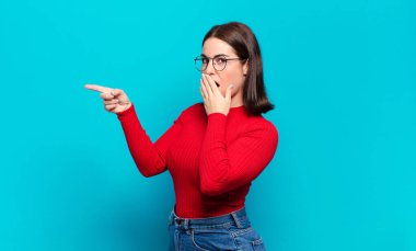 Genç, hoş, sıradan bir kadın kendini mutlu, şok olmuş ve şaşırmış hissediyor. Ağzını eliyle kapatıyor ve yan fotokopi alanını işaret ediyor.