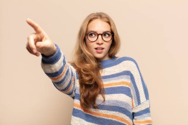 Şok olmuş ve şaşırmış hisseden sarışın bir kadın, şaşkınlık içinde, ağzı açık bir bakışla yukarı bakıyor ve onu işaret ediyor.