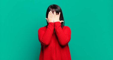 Artı boyda güzel bir kadın elleriyle yüzünü kapatıyor, parmaklarının arasından şaşkın bir ifadeyle yan tarafa bakıyor.