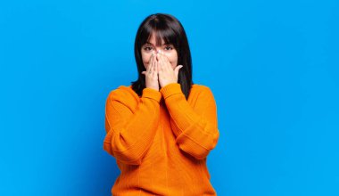 Artı boyunda güzel bir kadın mutlu, şanslı ve şaşkın görünüyor. İki eliyle ağzını kapatıyor.