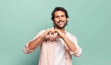 Genç, yakışıklı Hintli adam gülümsüyor ve mutlu, sevimli, romantik ve aşık, iki eliyle kalbi şekillendiriyor.