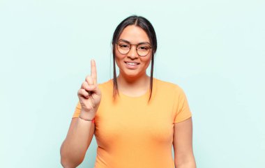 Latin bir kadın gülümsüyor ve arkadaş canlısı görünüyor, bir numarayı gösteriyor ya da ilk eli önde, geri sayıyor.