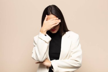 Genç İspanyol kadın stresli, utanmış ya da üzgün görünüyor, baş ağrısı var, yüzünü eliyle kapatıyor.