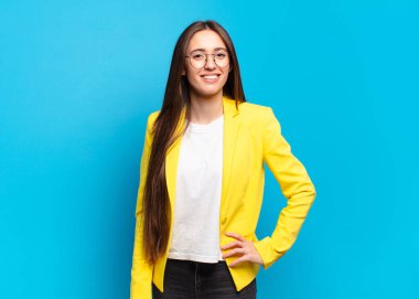 Genç ve güzel bir kadın mutlu bir şekilde gülümsüyor, eli kalçasında ve kendinden emin, pozitif, gururlu ve arkadaş canlısı.