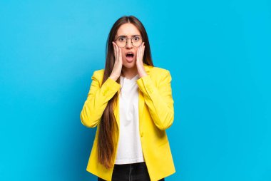 Genç ve güzel bir kadın şok olmuş ve korkmuş hissediyor, ağzı açık ve elleri yanaklarında korkmuş görünüyor.