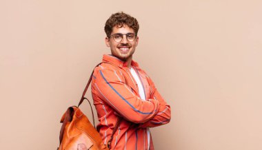 Genç adam kameraya çapraz kollarla gülümsüyor ve mutlu, kendinden emin, tatmin olmuş bir ifade, yan görüş. öğrenci konsepti
