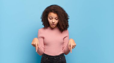 İki eliyle aşağıya doğru bakan ağzı açık bir kız. Şok olmuş, şaşırmış ve şaşırmış görünüyor.