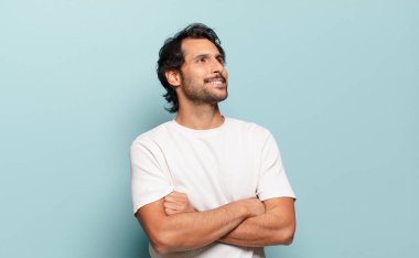 Genç ve yakışıklı Hintli adam mutlu, gururlu ve umutlu hissediyor, merak ediyor veya düşünüyor, çapraz kollarla boşluğu doldurmaya çalışıyor.