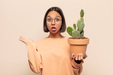 Genç Latin kadın şaşırmış ve şoka uğramış görünüyor, çenesi düşmüş, eli açık bir objeyi tutuyor.