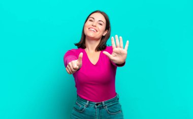 Genç, hoş, sıradan bir kadın gülümsüyor ve arkadaş canlısı görünüyor, altı ya da altıncı numarayı gösteriyor, eli önde, geriye doğru sayıyor.