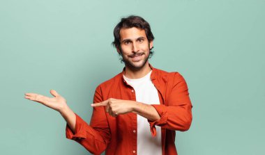 Genç, yakışıklı Hintli adam neşeyle gülümsüyor ve yan taraftaki fotokopi alanını işaret ediyor, bir nesneyi gösteriyor ya da reklamını yapıyor.