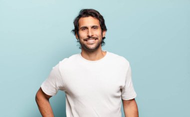 Genç ve yakışıklı Hintli adam mutlu bir şekilde gülümsüyor, eli kalçasında ve kendinden emin, olumlu, gururlu ve arkadaş canlısı.