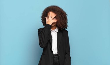 Şok olmuş, korkmuş ya da dehşete düşmüş, yüzü elle kaplanmış ve parmaklarının arasından gözetleyen bir afro genç. mizahi iş konsepti