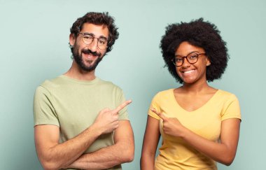multiracial couple of friends smiling cheerfully, feeling happy and pointing to the side and upwards, showing object in copy space clipart