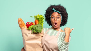 Genç, güzel, afro bir kadın inançsızlıktan şaşkın şaşkın bakıyor, yan taraftaki objeyi işaret ediyor ve diyor ki vay be, inanılmaz ve elinde sebze torbası var.