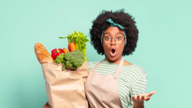 Genç, güzel, afro bir kadın ağzı açık ve şaşırmış, şok olmuş ve hayrete düşmüş. İnanılmaz bir sürpriz ve elinde bir sebze torbası var.