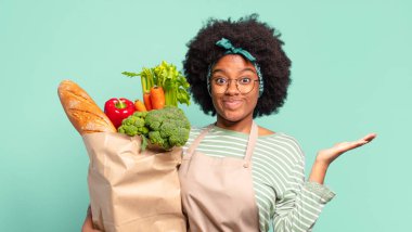 Genç, güzel afro bir kadın kafası karışık ve kafası karışık, başını kaşıyor ve yan tarafa bakıyor ve elinde bir sebze torbası tutuyor.