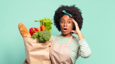 Genç, güzel bir afro kadın kendini stresli, endişeli, endişeli ya da korkmuş hissediyor. Elleri başının üstünde. Yanlışlıkla panikliyor ve elinde bir sebze torbası tutuyor.