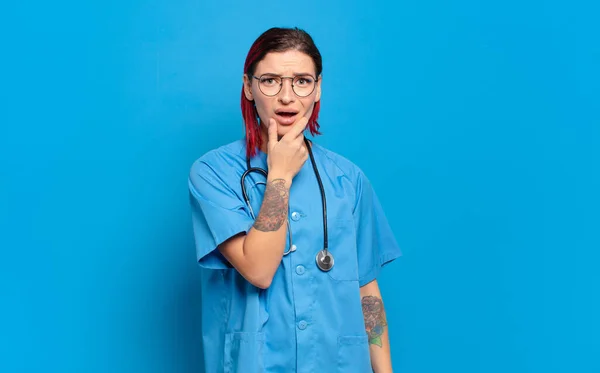 口や目を大きく開いて顎を手に若い魅力的な赤い髪の女性は 不快なショックを感じ 何やすごいと言う 病院看護師のコンセプト — ストック写真