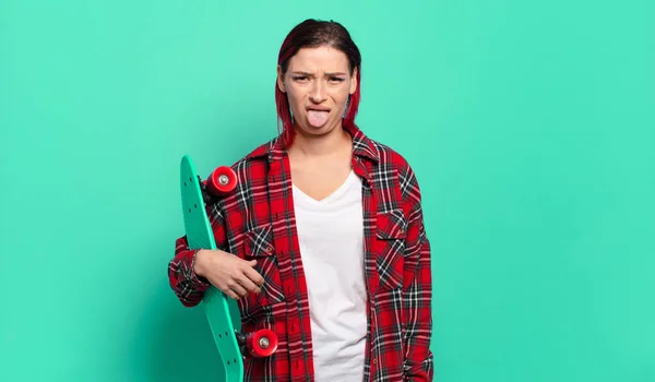 若い魅力的な赤い髪の女性はうんざりし いらいらし 舌を突き出し 何か厄介で幸運を嫌い スケートボードを保持します — ストック写真