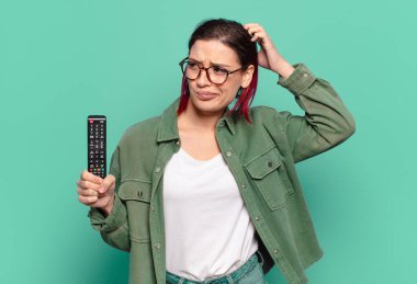 Genç ve çekici kızıl saçlı kadın kafası karışık, kafasını kaşıyor ve yan tarafa bakıyor ve elinde uzaktan kumandayla televizyonu tutuyor.