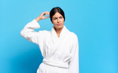 Genç İspanyol kadın şaşkın ve şaşkın hissediyor, başını kaşıyor ve yana bakıyor. Bornoz konsepti