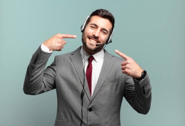 Genç işadamı güvenle gülümseyerek kendi geniş gülümsemesine olumlu, rahat, tatmin olmuş tele pazarlama konseptine işaret ediyor.