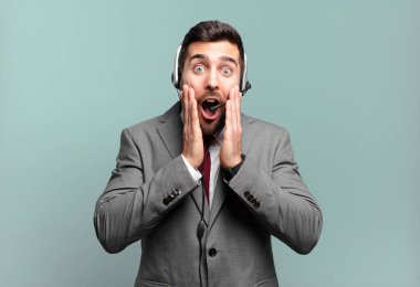 Genç işadamı şok olmuş ve korkmuş hissediyor, ağzı açık ve elleri yanaklarında tele pazarlama konseptiyle korkmuş görünüyor.